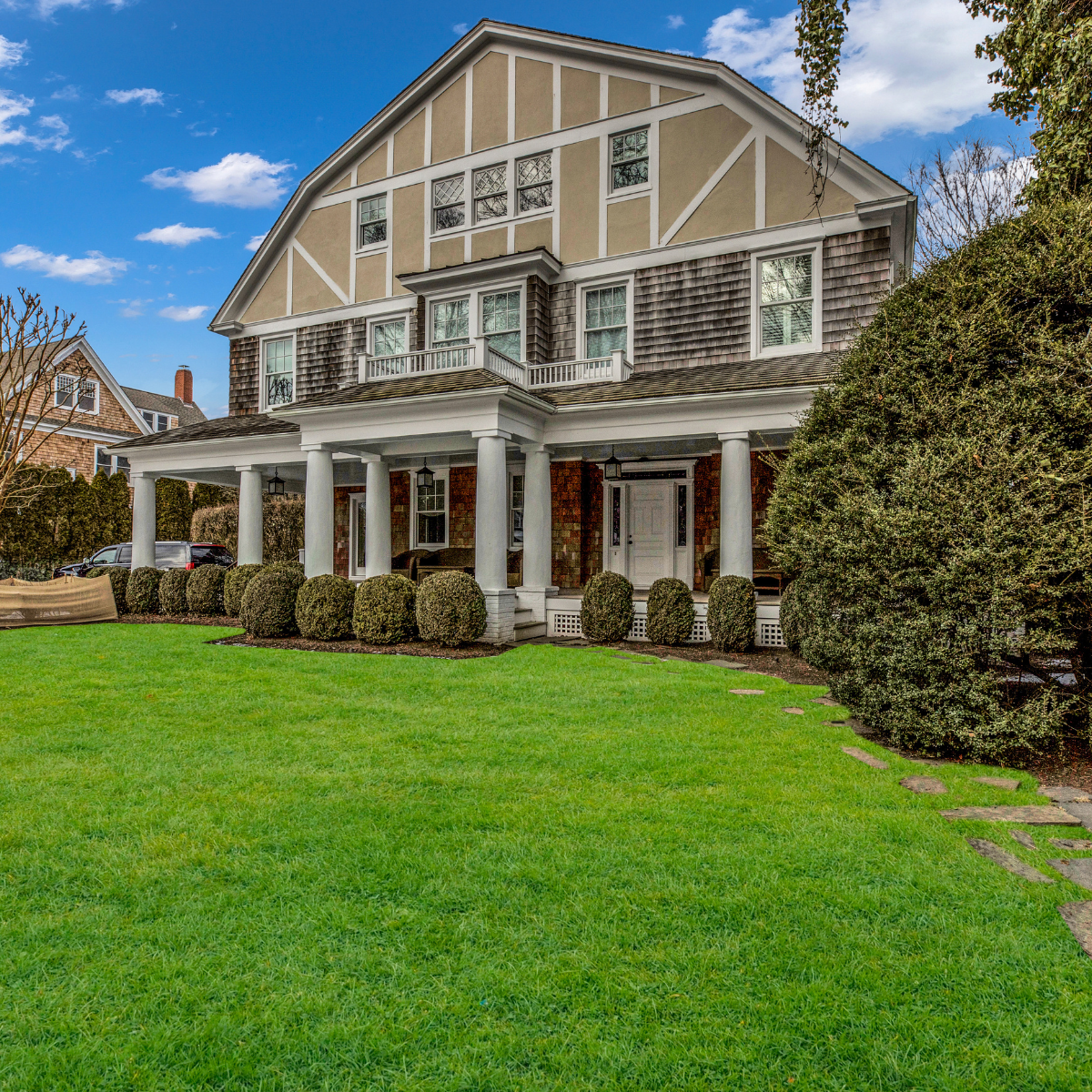 Two-story house with a gambrel roof, a large front porch supported by white columns, manicured lawn, and neatly trimmed bushes under a partly cloudy sky.