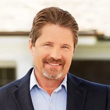 A middle-aged man with short brown hair, a trimmed beard, and mustache, wearing a blue shirt and dark blazer, smiling outdoors.
