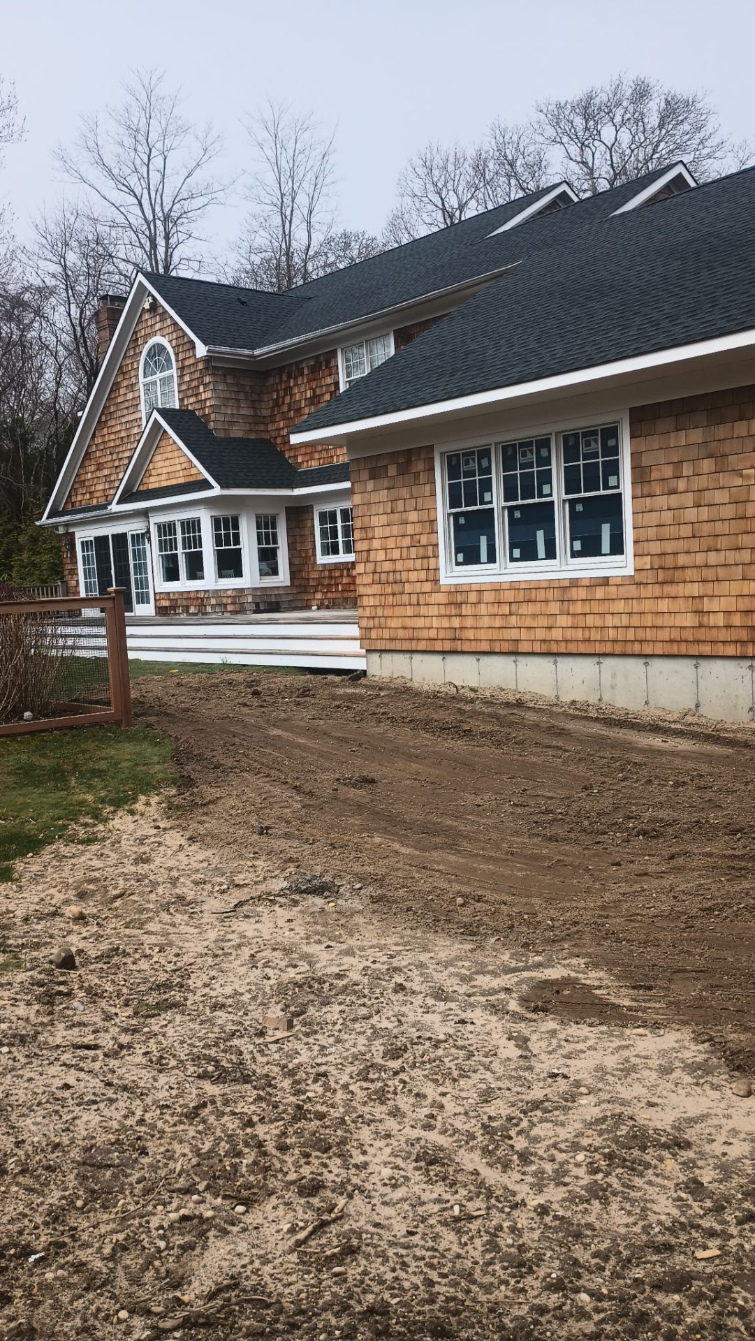 A house with wood shingle siding and multiple windows, surrounded by bare trees, with a dirt driveway in the foreground.