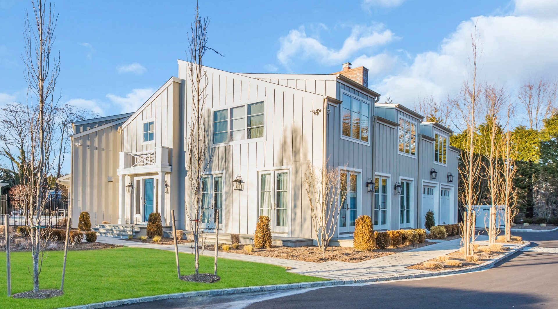 Modern two-story house with large windows, light gray siding, minimal landscaping, and a paved walkway under a blue sky.